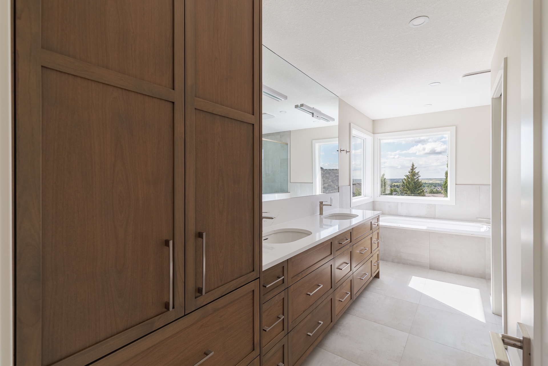 Bright modern bathroom with wooden cabinets, double sink vanity, large mirror, and a bathtub by a window showing an outdoor view by Marvel Cabinetry.