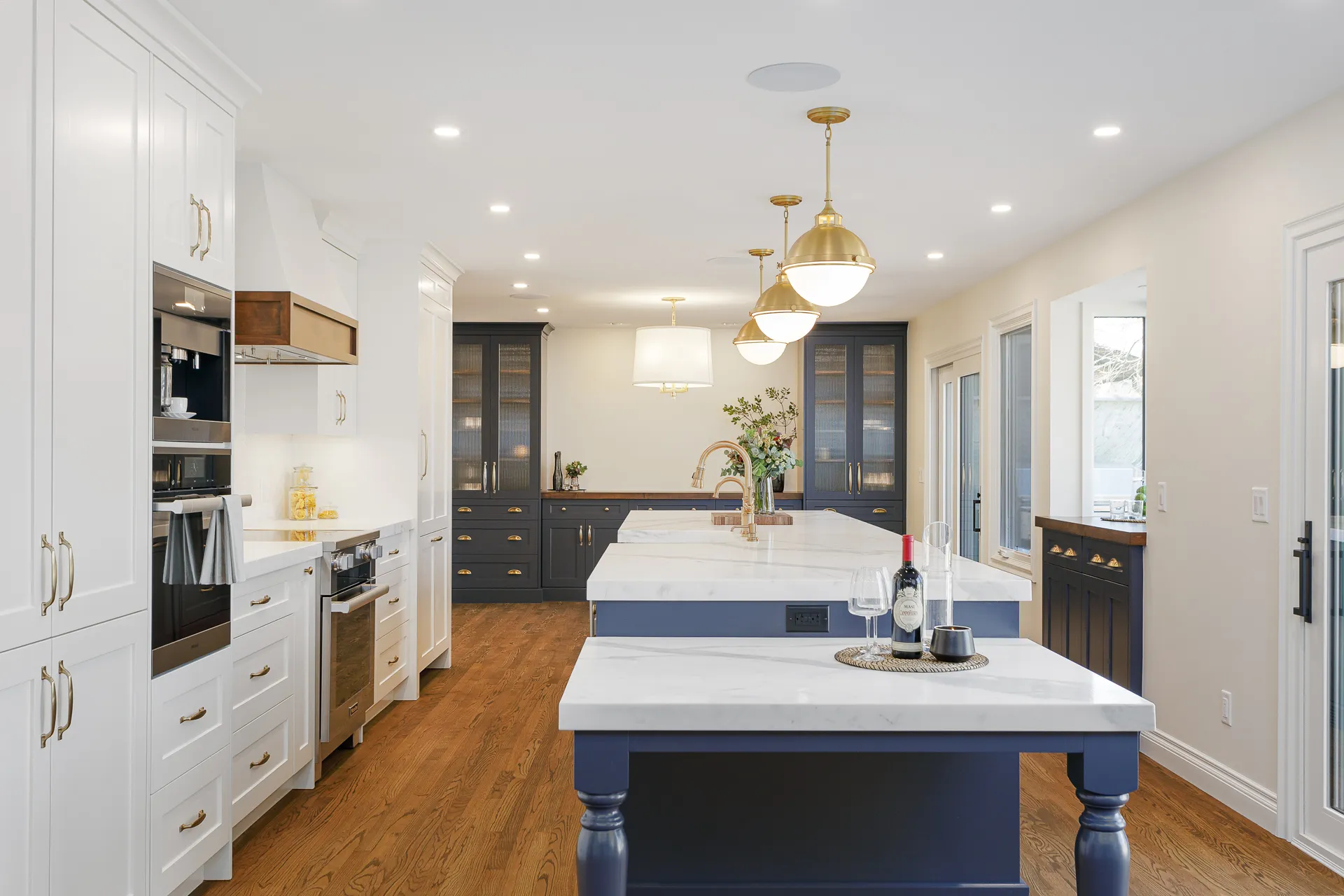 Modern kitchen with white and navy blue cabinetry, large marble island with gold fixtures, hardwood floors, and pendant lights by Marvel Cabinetry.