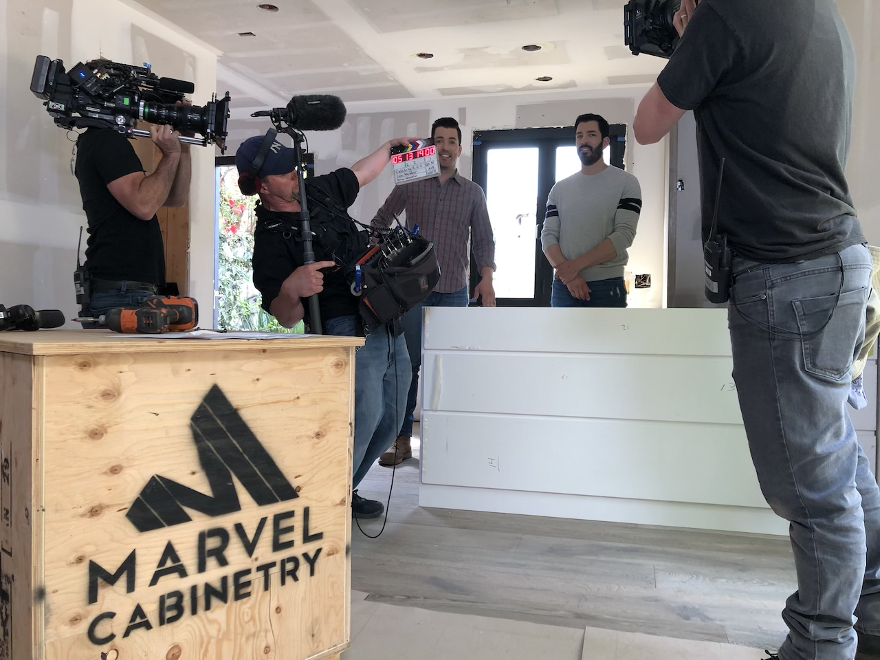 Two men standing behind a white kitchen island being filmed by two camera operators in a room with Marvel Cabinetry branding on a wooden crate during a shoot for HGTV's The Property Brothers Celebrity I.O.U.