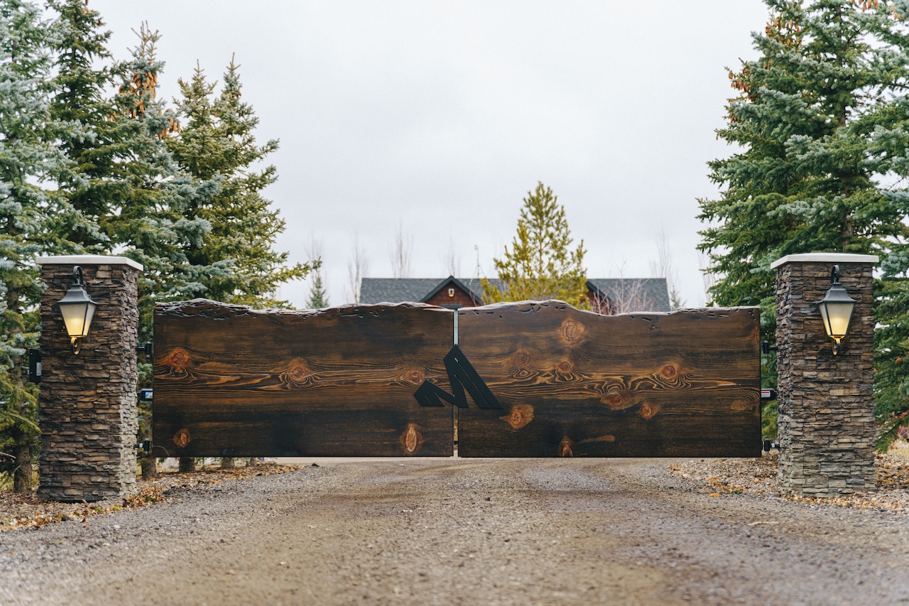 Marvel Cabinetry & Renovations entrance. Rustic wooden gate with a black metal emblem between two stone pillars with lamps on a gravel driveway surrounded by pine trees.