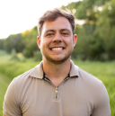 Smiling young man in a tan polo shirt standing outdoors with blurred green trees in the background.
