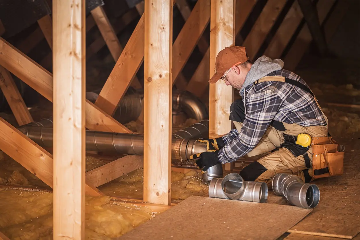 A male HVAC technician wearing work gear, a plaid shirt, and a cap is crouched down in a dark attic space. He is connecting metal ductwork fittings, surrounded by wooden framing and yellow fiberglass insulation on the floor. Several loose duct fittings are visible on a piece of oriented strand board (OSB) in the foreground.