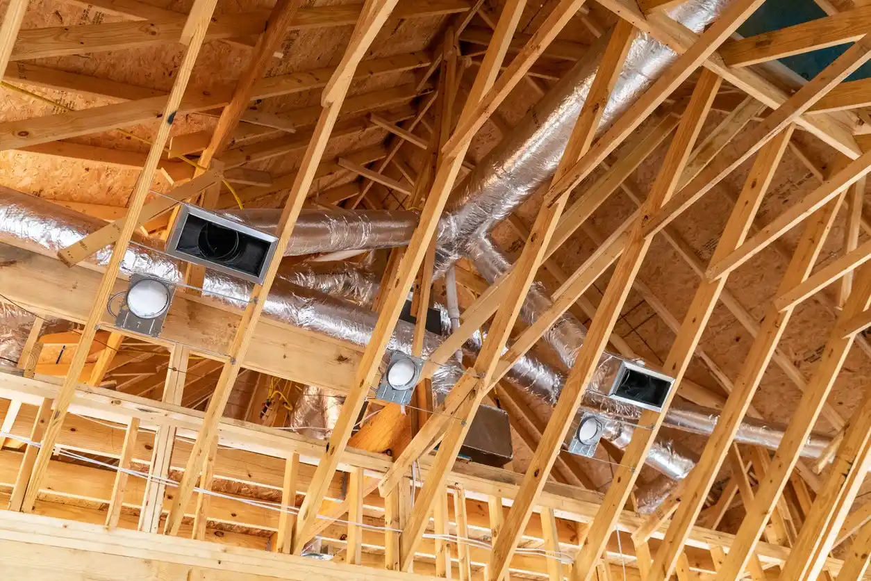 A low-angle view looking up into the exposed wooden frame and rafters of a newly constructed roof. Several insulated flexible air ducts wrapped in silver foil are run between the rafters, and four metal duct boxes for vents or recessed lights are installed and connected to the ductwork.