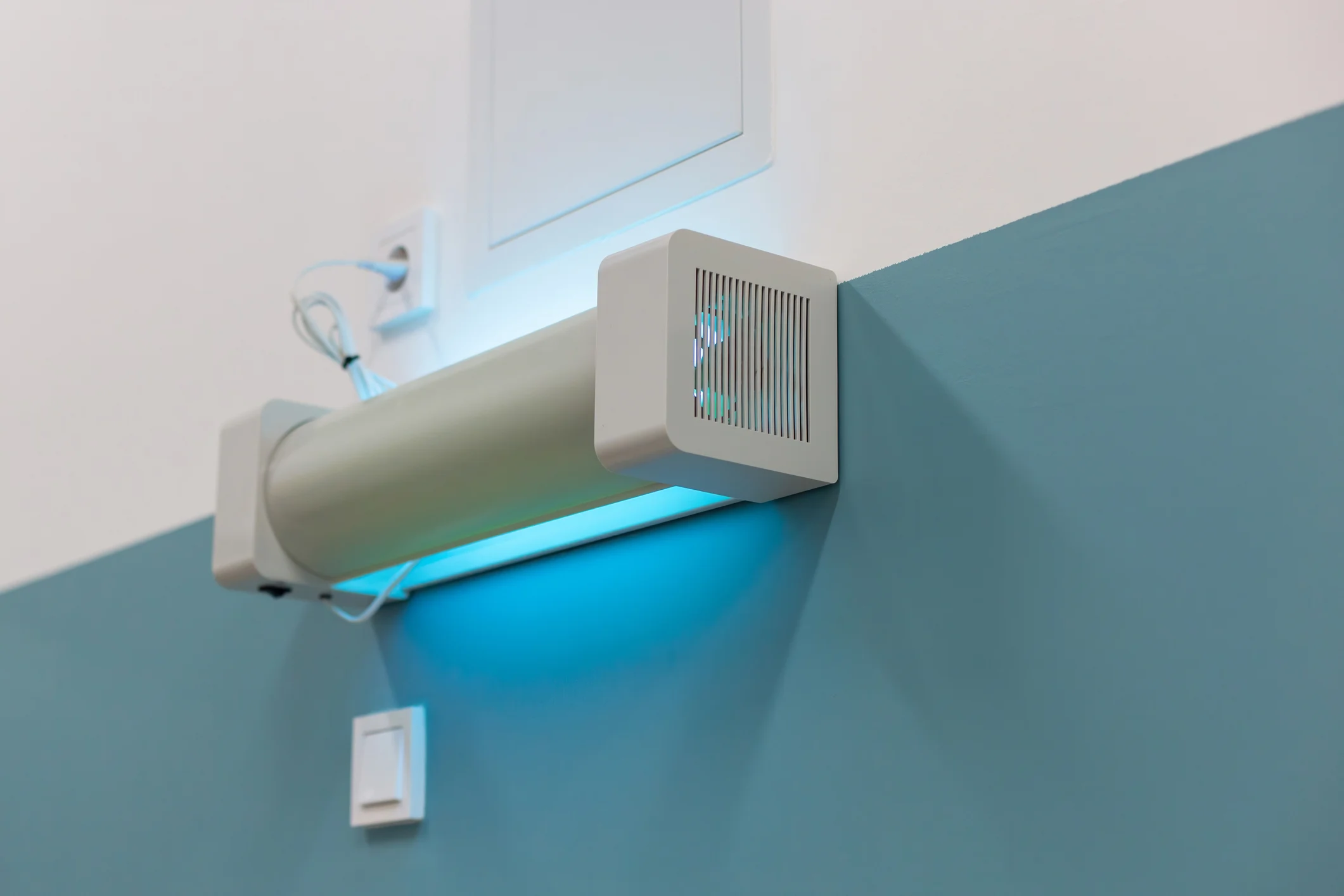 A white, cylindrical UV air purifier or germicidal light is mounted horizontally high on a wall, with its ends enclosed by square white boxes that feature vertical vents. The fixture emits a strong blue-cyan light and is installed where a light blue wall meets a white ceiling. Wires are connected near a power outlet above the unit.
