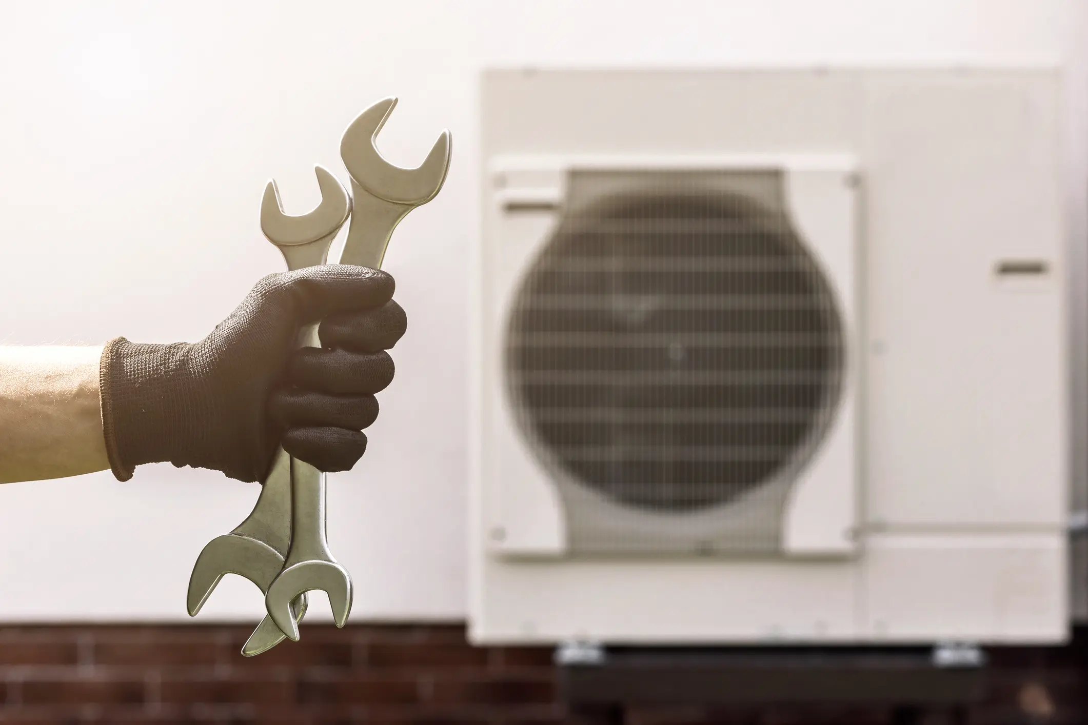 A person's hand, wearing a black work glove, is holding two large silver wrenches against a bright background. The background is blurred, showing a white outdoor air conditioning or heat pump unit with a circular fan grille.