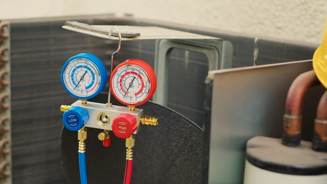A close-up shot of an HVAC technician's manifold gauge set, with blue (low pressure) and red (high pressure) gauges, connected to the copper refrigerant lines of an outdoor air conditioning or heat pump condenser unit. The unit's dark aluminum fins and a cylindrical component are visible in the background.