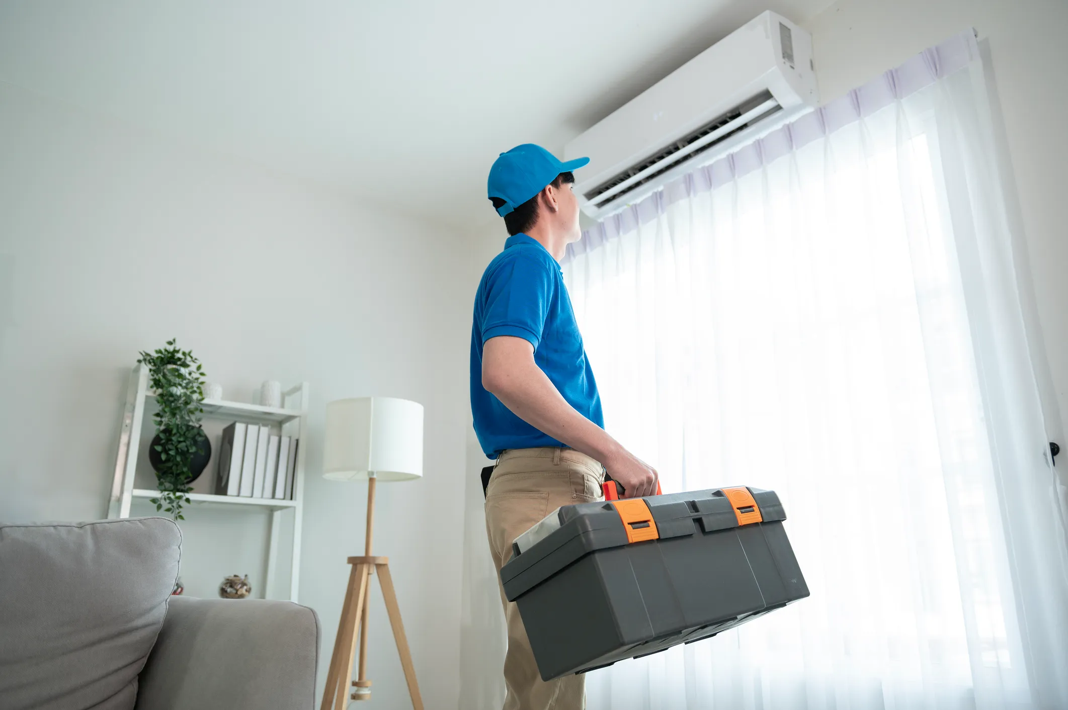 HVAC technician carrying a toolbox and looking up at an indoor air conditioner in a living room.