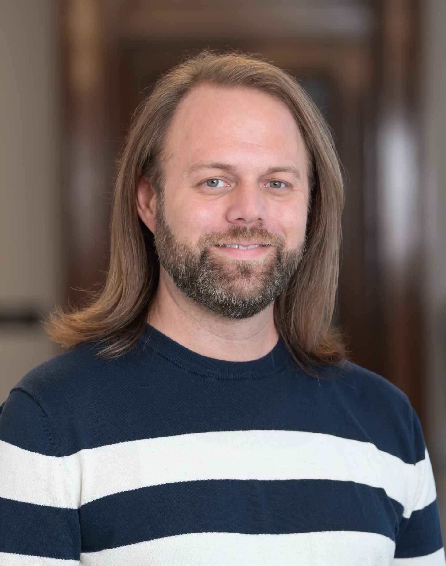 Tony Baker - smiling man with shoulder-length light brown hair and beard wearing a navy blue and white striped sweater.