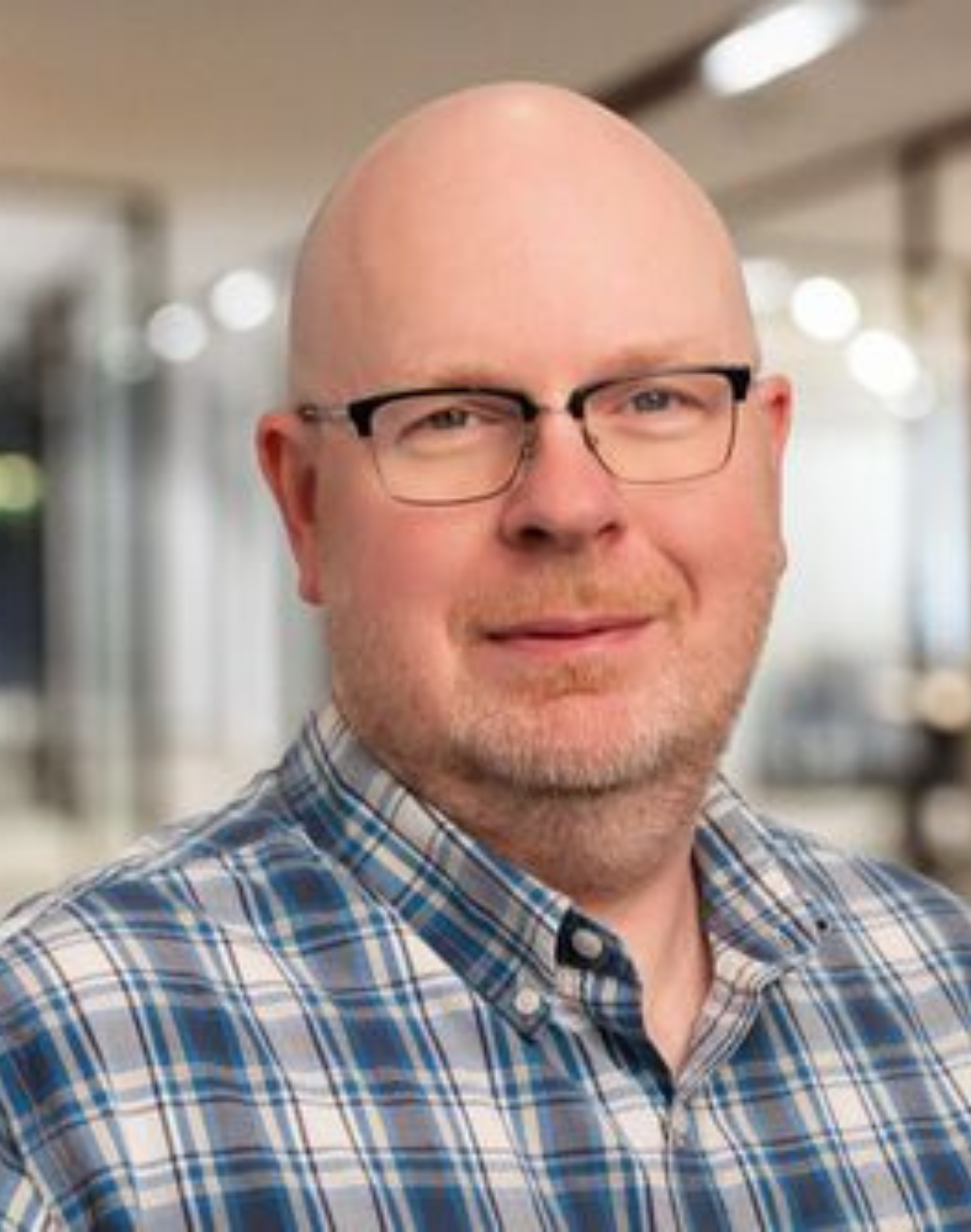 Scott Erickson - middle-aged bald man with glasses wearing a blue and white plaid shirt, smiling slightly in an office setting.