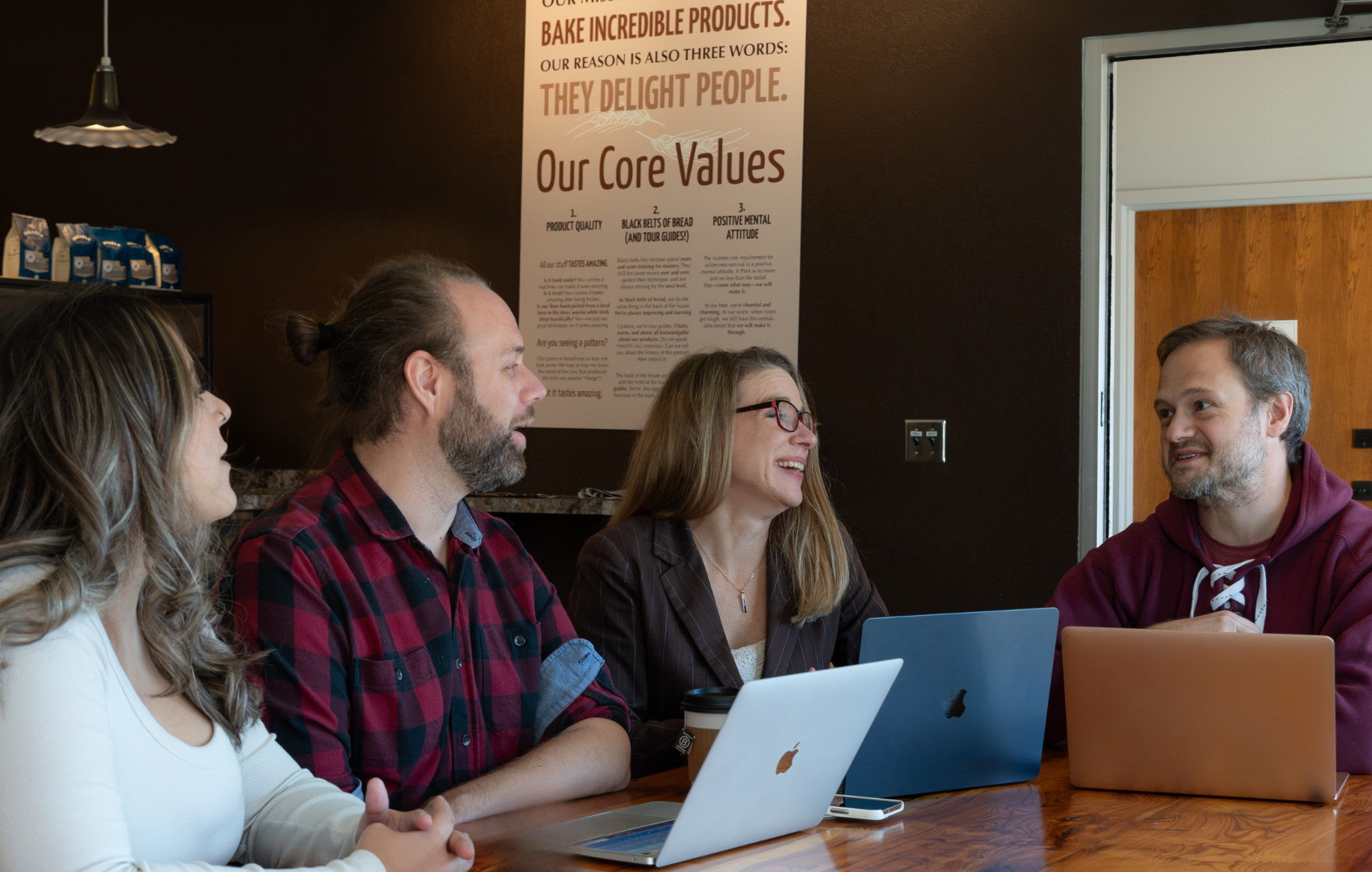 Four people in a meeting room with laptops, engaged in a discussion and smiling.