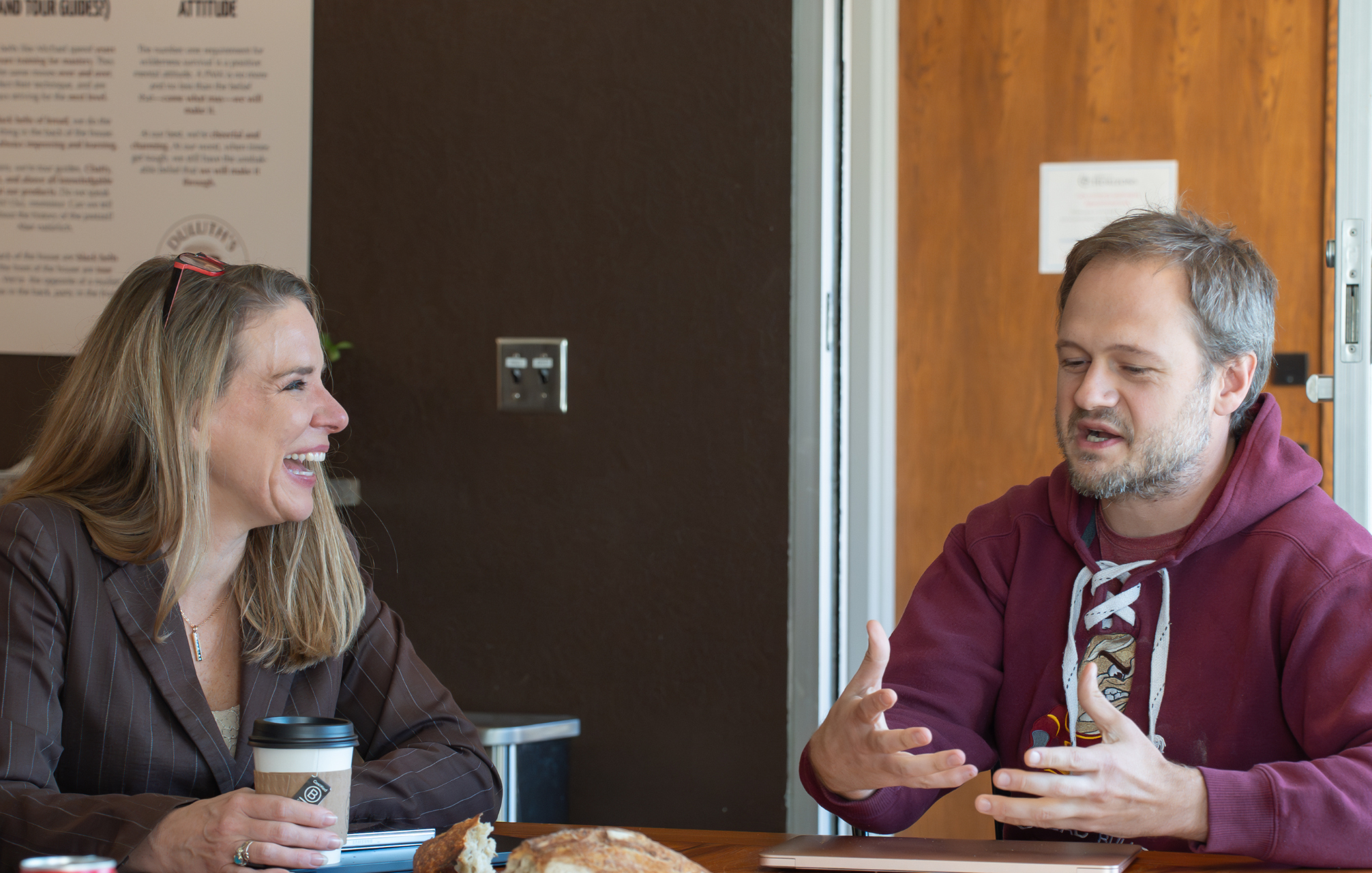 Two adults seated at a table having a conversation, one woman laughing while holding a coffee cup and a man speaking with hand gestures.