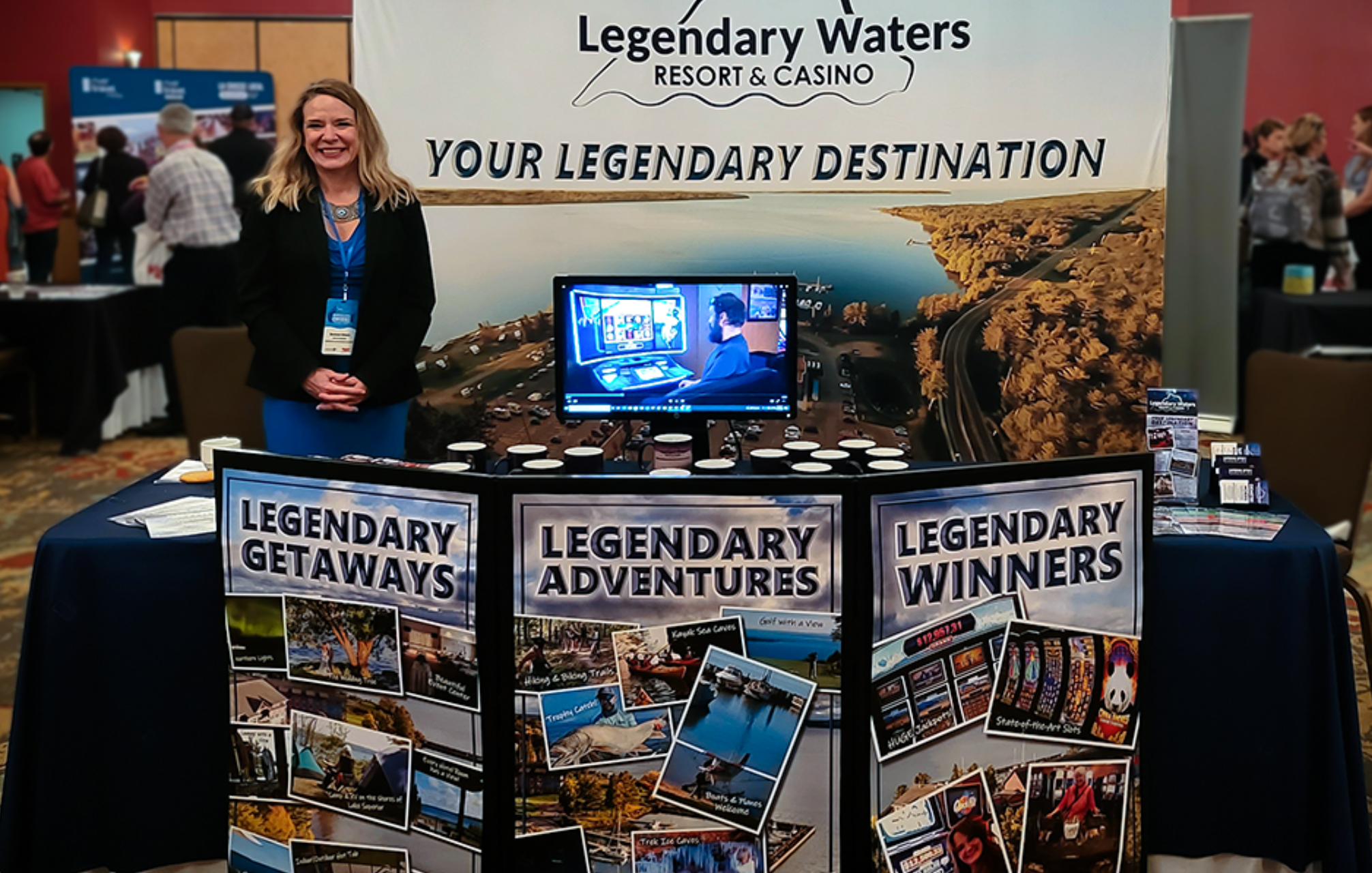 Exhibition booth for Legendary Waters Resort & Casino featuring displays titled Legendary Getaways, Legendary Adventures, and Legendary Winners with a smiling woman standing behind the table.