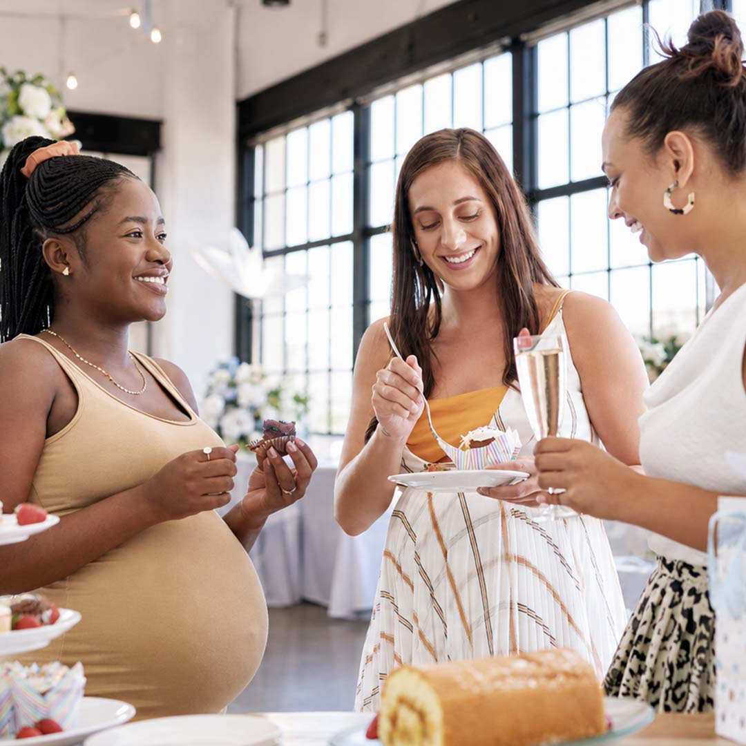 Three women smiling and enjoying cupcakes and drinks at a bright indoor celebration.