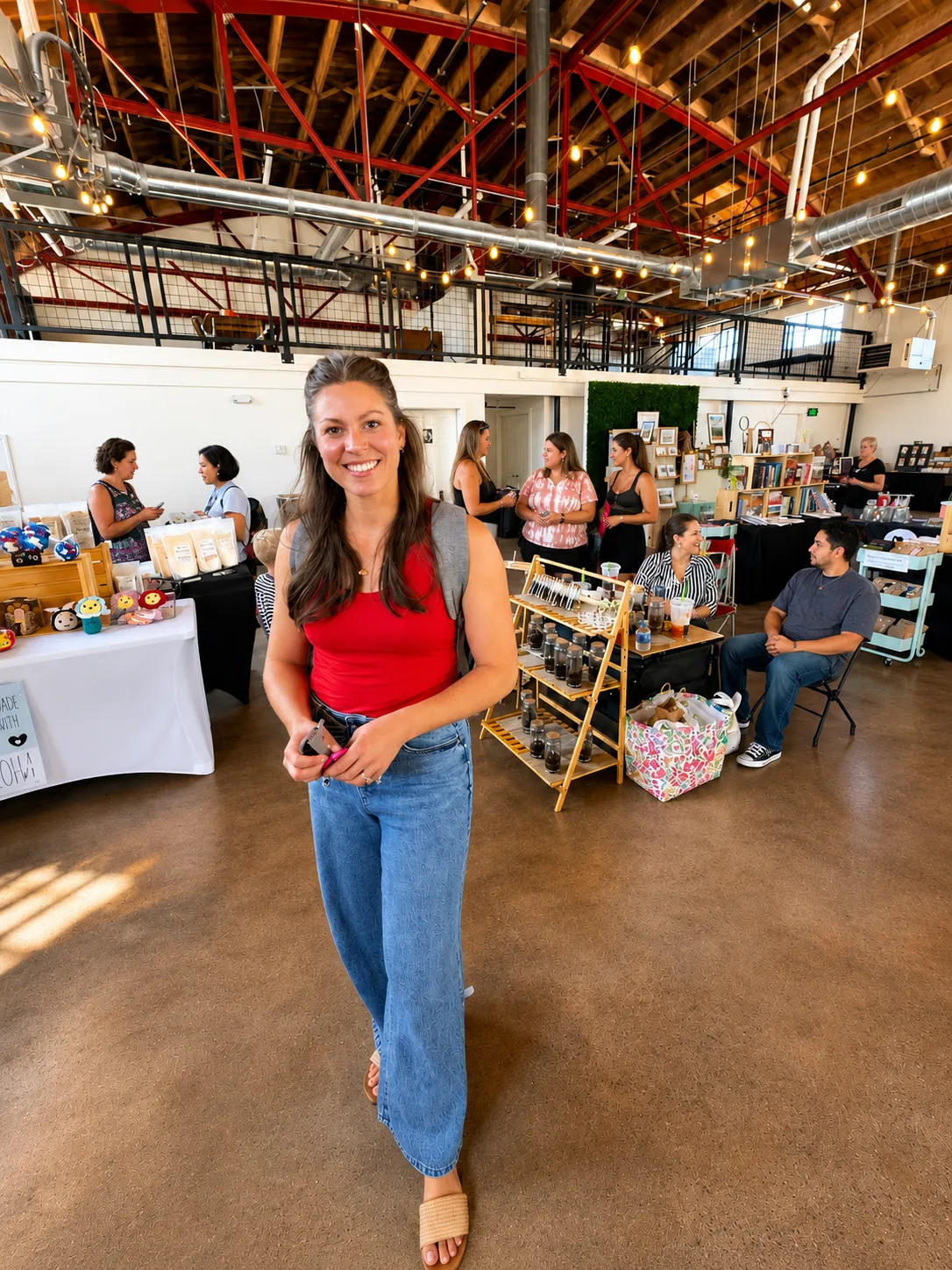 Smiling woman in red top and jeans walking inside a lively indoor market with booths and people.