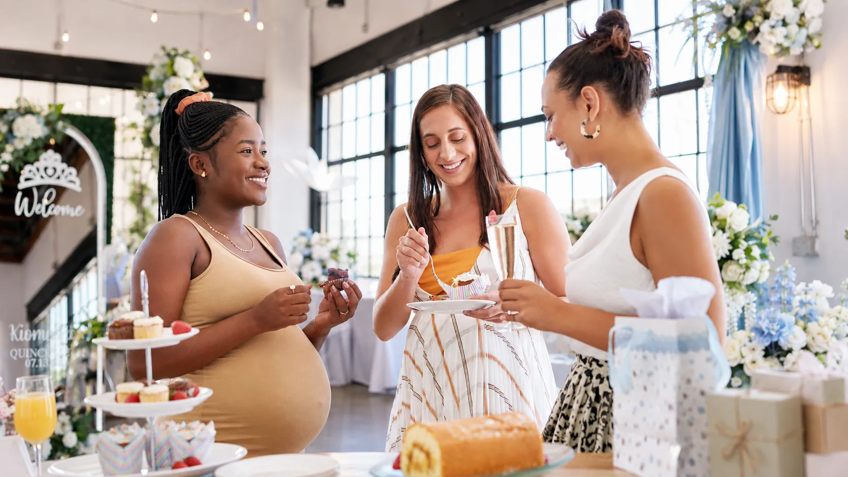 Three women enjoying cupcakes and drinks at a decorated baby shower with gifts and flowers.