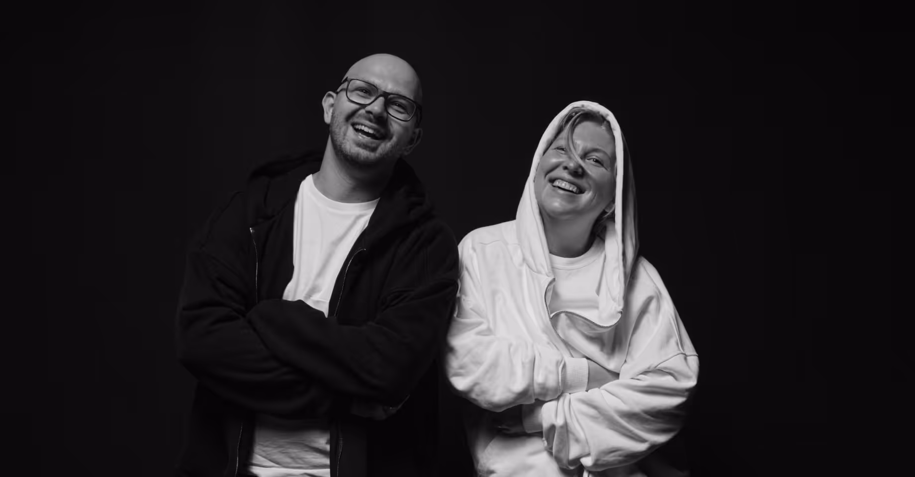 Smiling man and woman standing with arms crossed against a black background in black and white photo.