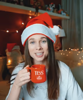 Woman wearing a Santa hat holding a red TESS mug with Christmas lights in background.