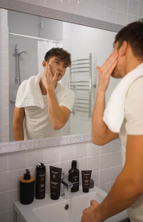 Young man with a towel around neck applying skincare cream in front of bathroom mirror.