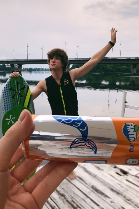 Person in life vest holding a wakeboard on a dock with a can of Fanta in the foreground near a river bridge.