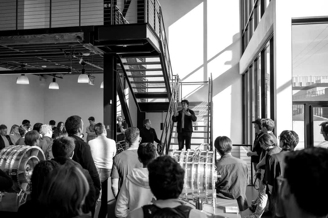 A speaker addresses a crowd of engineers and operators in a sunlit hardware lab