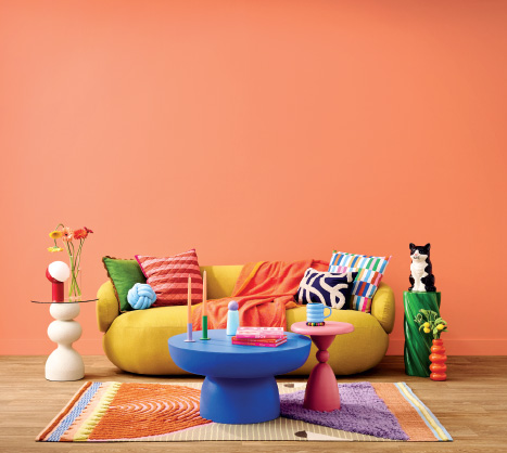 A vibrant maximalist living room featuring a curved yellow sofa with eclectic throw pillows in stripes and geometric patterns. In front of it is a bold cobalt blue pedestal coffee table and a smaller pink side table. The space is anchored by a colourful abstract rug with orange and purple tones, set against a solid Sico paint orange wall. Playful decor includes a white sculptural side table, a green twist pedestal with a tuxedo cat figurine, and colourful tapered candles.