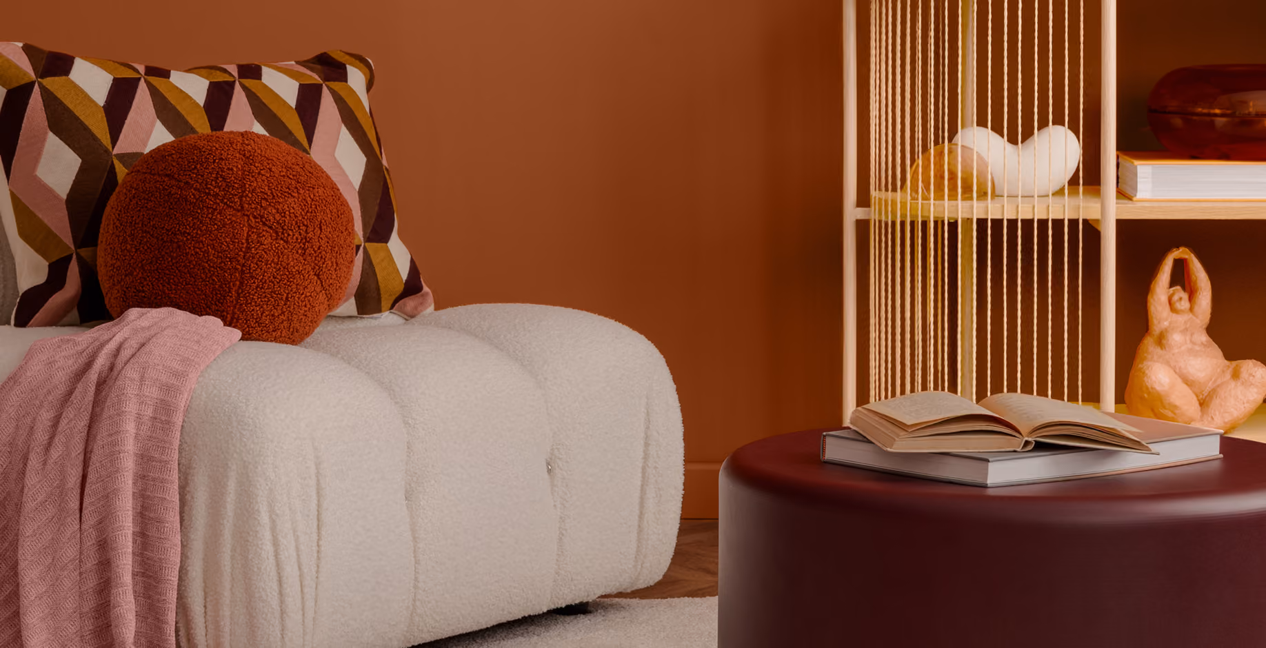 A close-up of a cozy living room corner with a terracotta wall, a white bouclé sofa with decorative pillows, and an open book resting on a dark burgundy ottoman