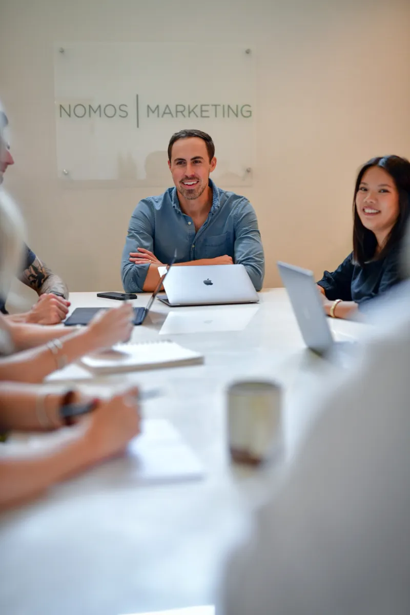 A man and woman sit at the head of a white conference table, engaged in conversation with their team during a meeting.
