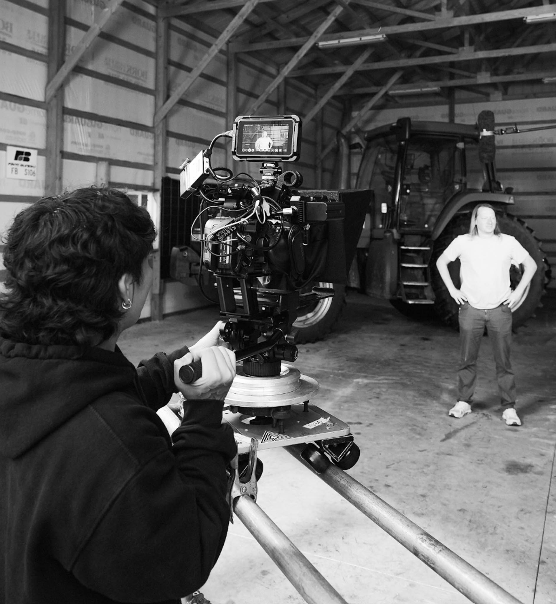 Person operating a professional video camera on a dolly track filming another person standing in front of a large tractor inside a barn.
