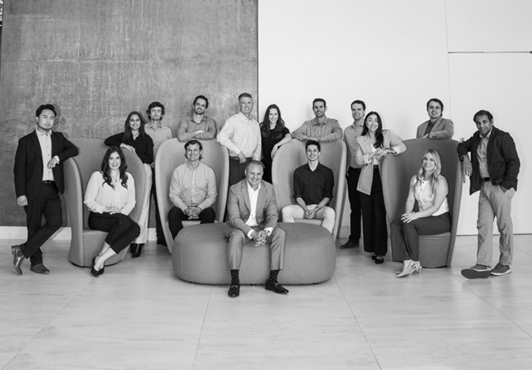 Black and white photo of a diverse group of 15 men and women, some seated on modern chairs and ottomans and others standing, posing in a bright, minimalist room.