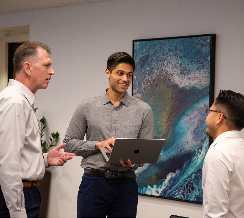 Three men in business casual attire having a discussion in an office, one holding a laptop and smiling.