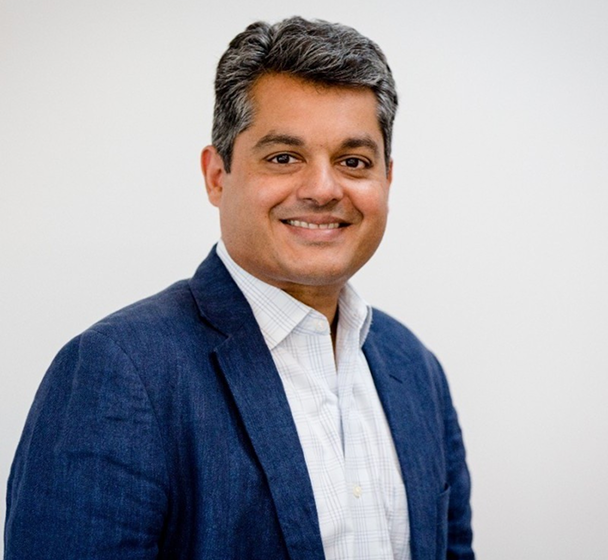 Smiling man with short black and gray hair wearing a blue blazer and white checkered shirt, standing against a plain white background.