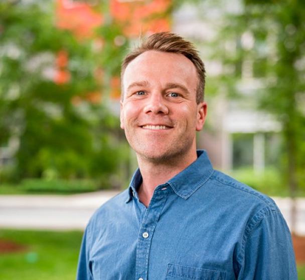Smiling man in a blue button-up shirt standing outdoors with blurred greenery and buildings in the background.