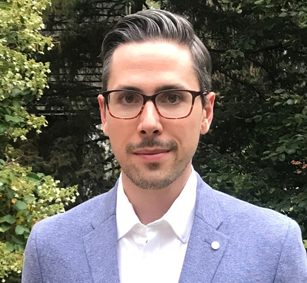Man with dark hair and glasses wearing a light blue blazer and white shirt standing in front of green trees.