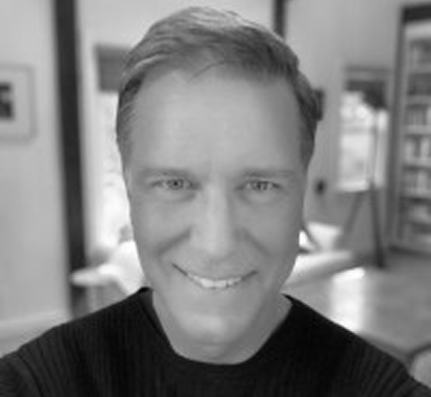 Black and white close-up photo of a smiling man with short hair wearing a dark sweater in a home setting.