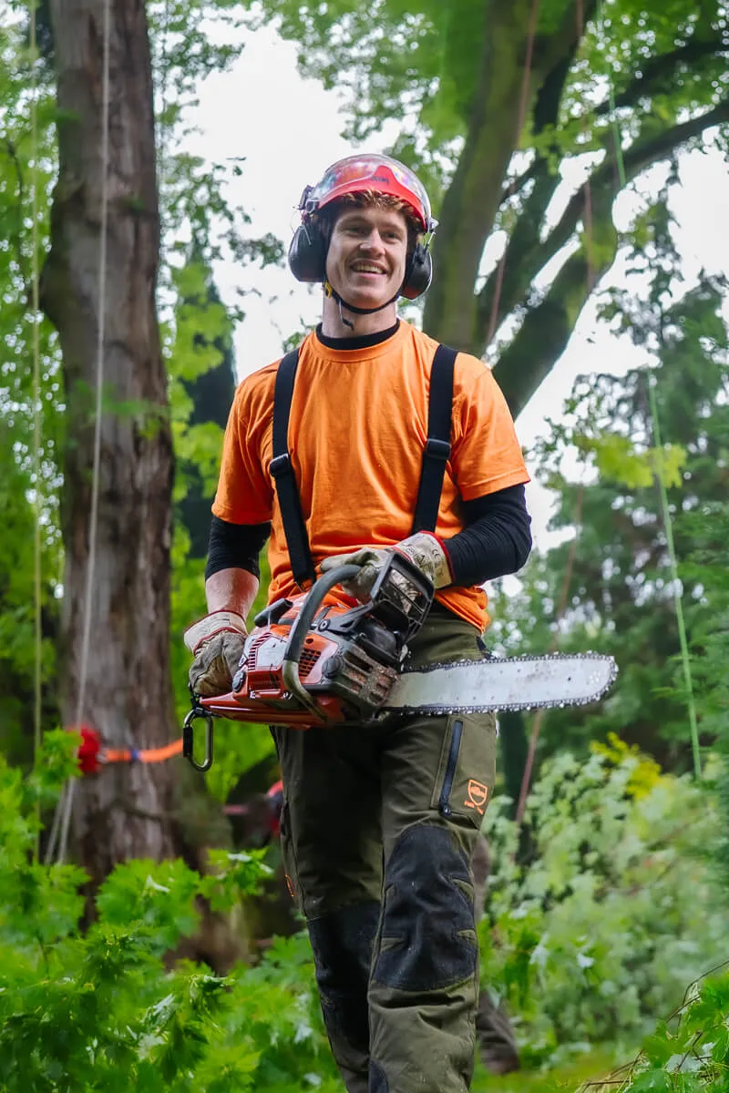 Smiling man in safety gear holding a chainsaw in a forest setting.