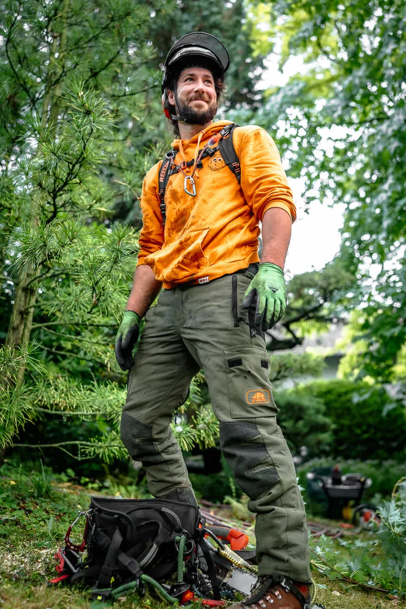 Man in orange hoodie, green gloves, and safety helmet standing outdoors with chainsaw and gear on ground.