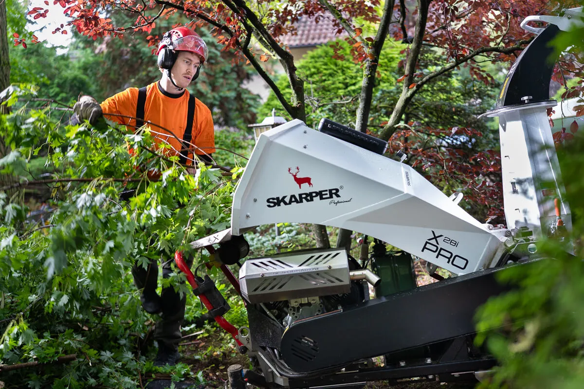 Worker wearing orange shirt and red helmet feeding tree branches into a white SKARPER wood chipper in a garden.