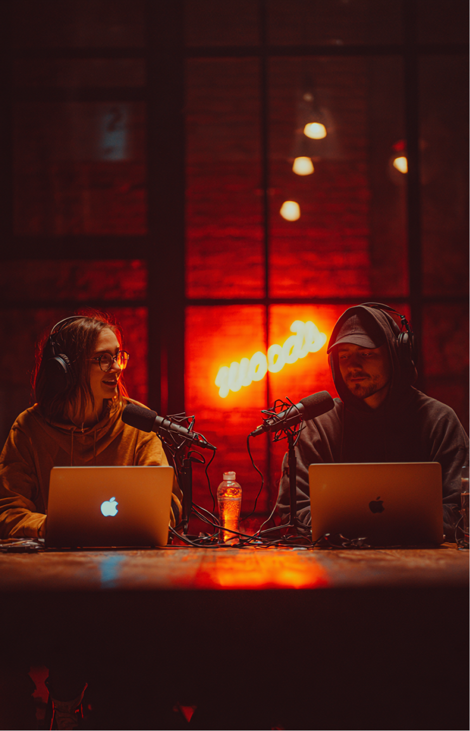 Two podcasters with headphones and laptops recording in a dimly lit studio with a neon sign saying 'moods' in the background.