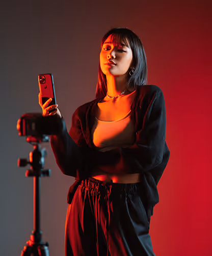 Young woman with long dark hair holding a smartphone and posing for a camera on a tripod with dramatic red and gray lighting.
