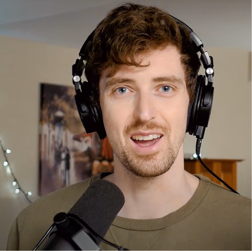 Young man with curly brown hair wearing black headphones speaking into a microphone indoors.