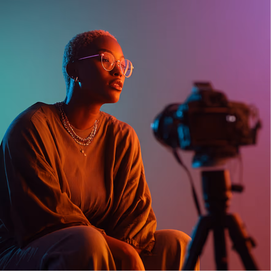 Person with short hair and glasses sitting in front of a camera on a tripod, illuminated by warm and cool lighting.