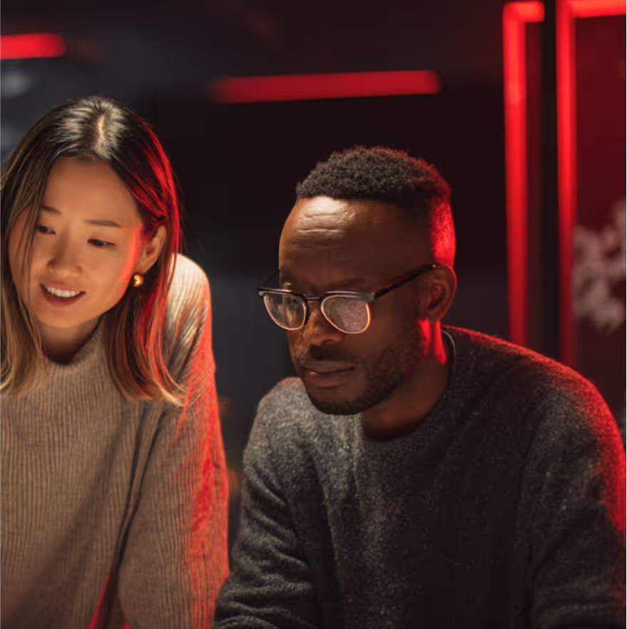 A woman and a man wearing glasses focused on a task under warm red lighting.