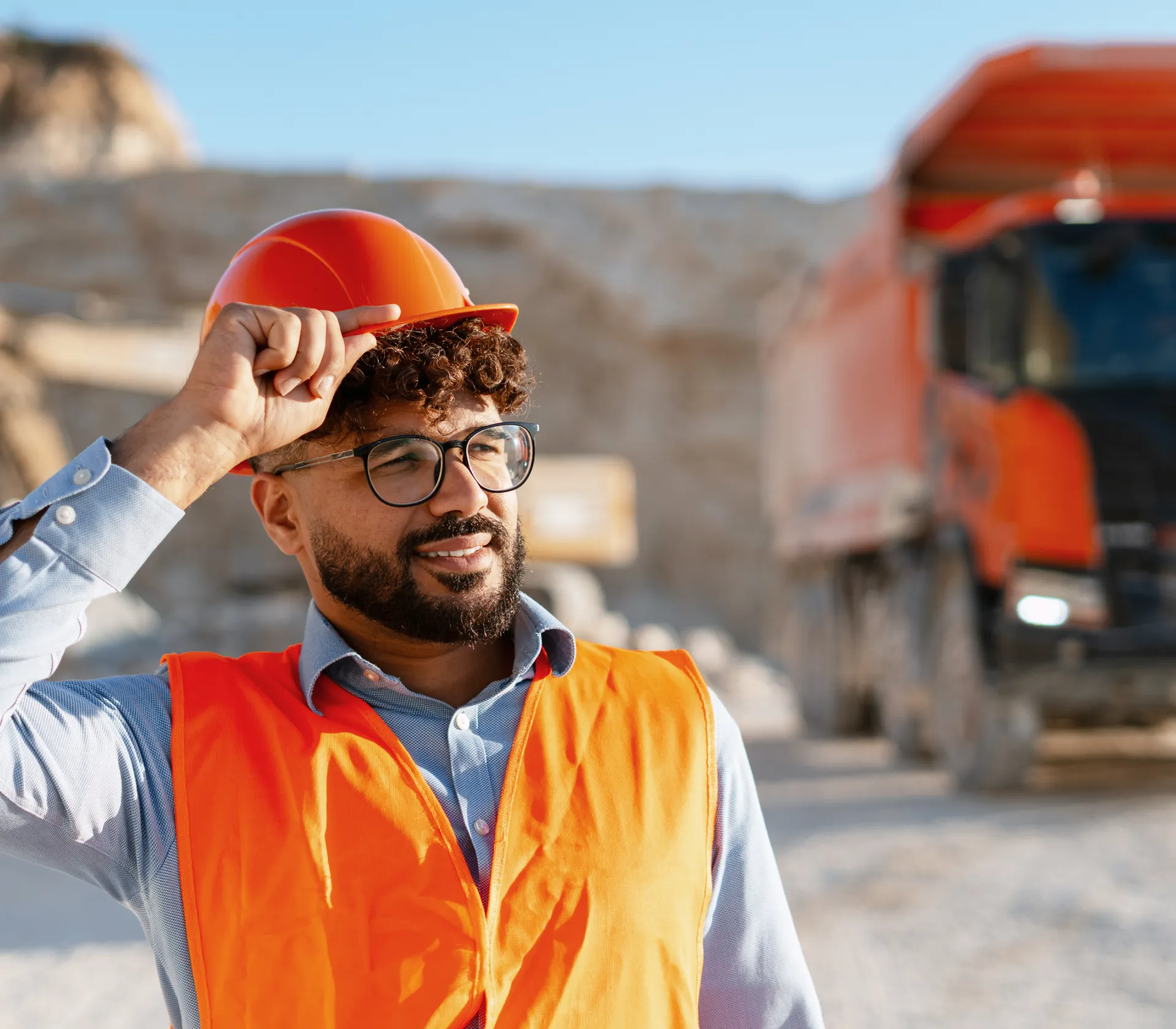 Smiling male construction worker wearing orange safety vest and hard hat, standing at a mining site with large orange mining truck in the background.
