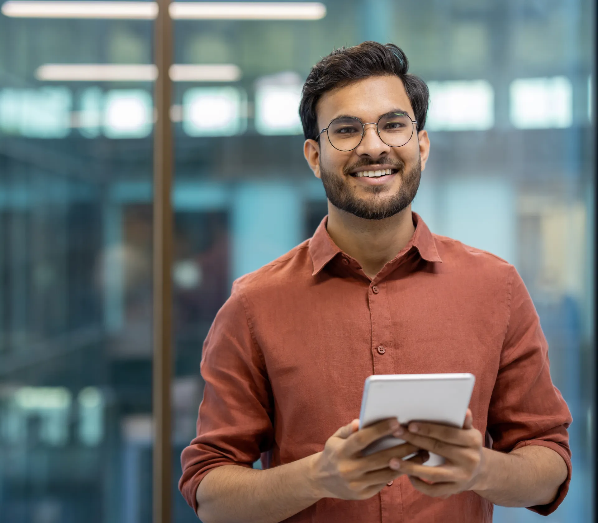 Smiling man wearing glasses and a rust-colored shirt holding a digital tablet in a modern office.