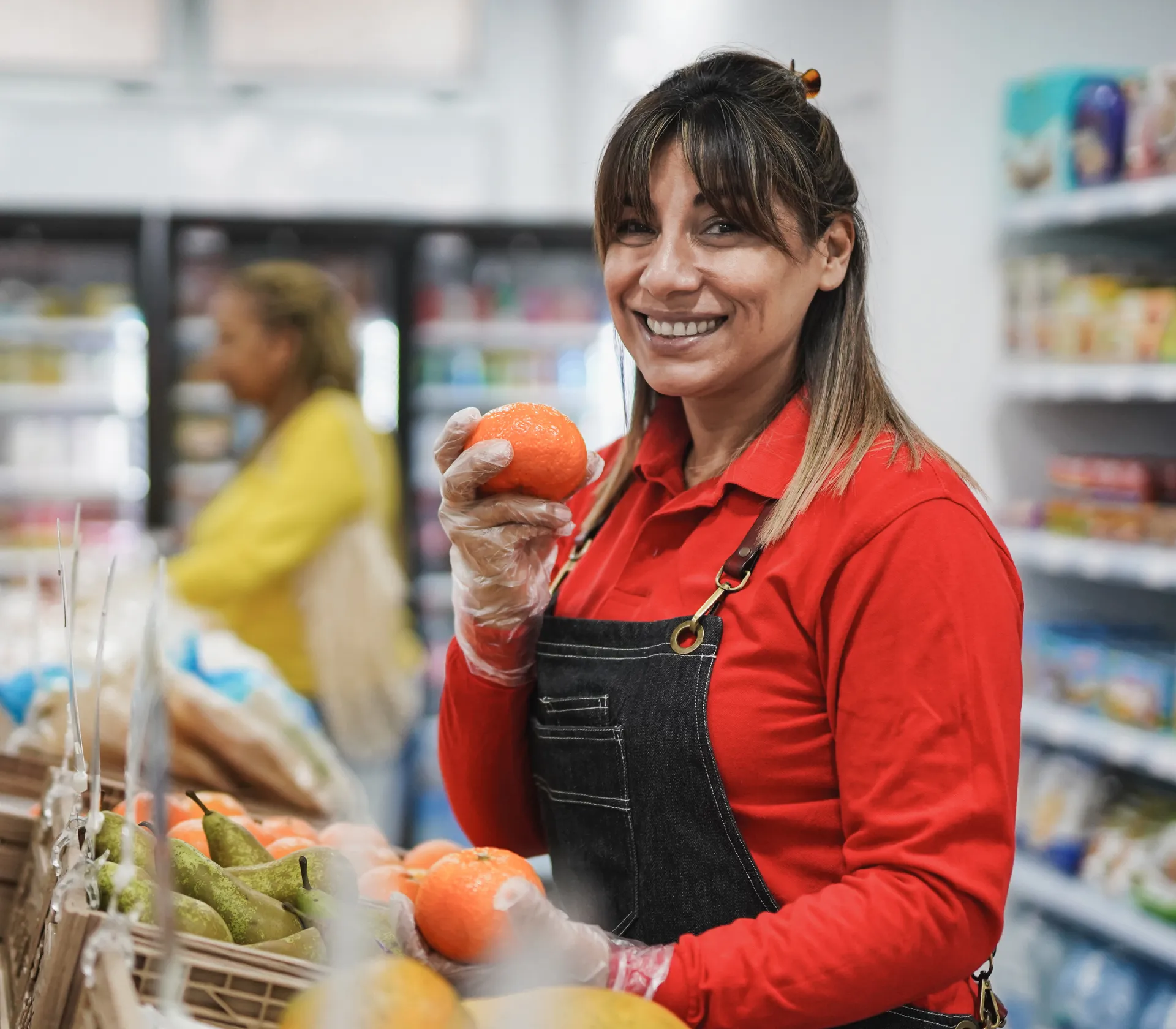 Smiling grocery store worker in red shirt and apron holding an orange among fresh produce.