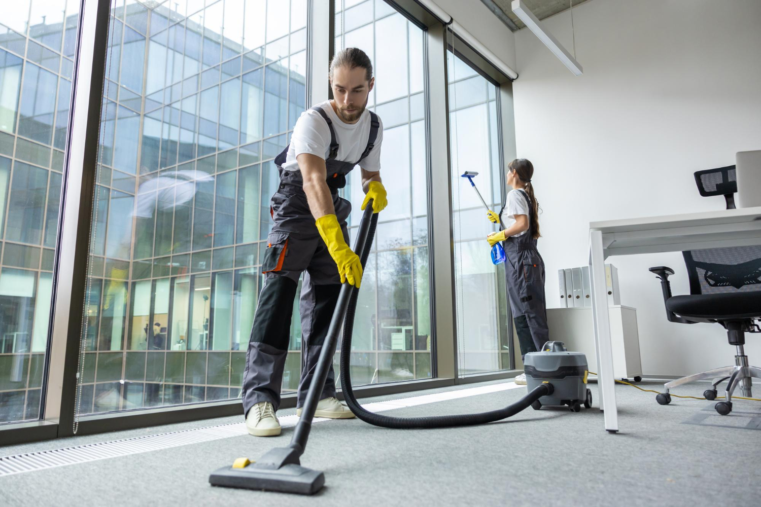 Two janitors wearing yellow gloves cleaning a modern office with large windows, one vacuuming carpet and the other cleaning windows.