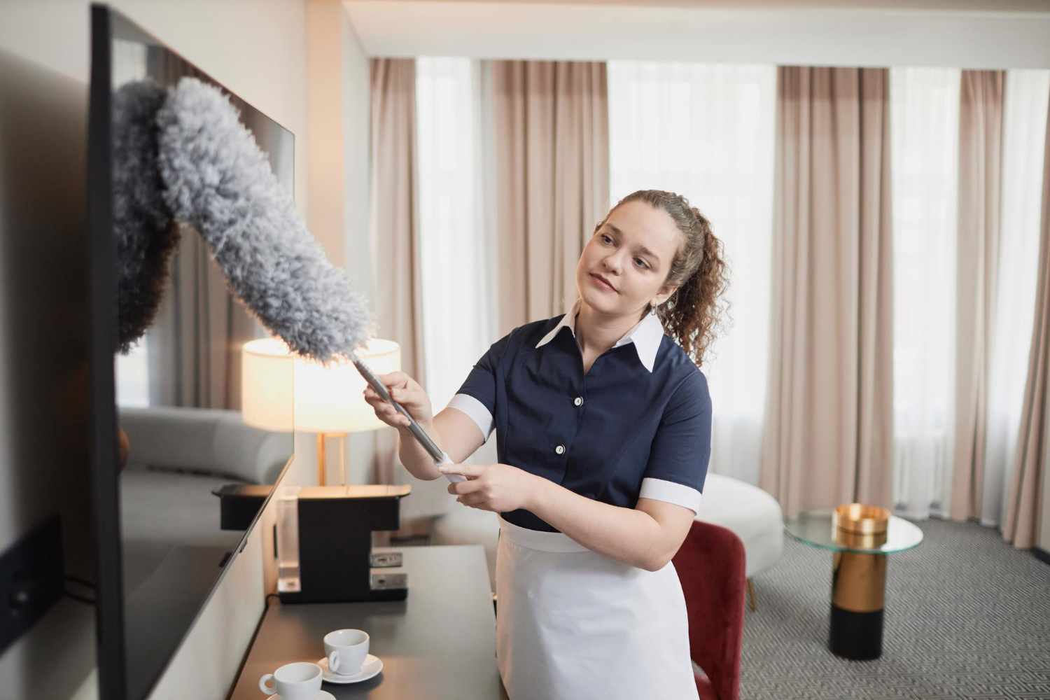 Housekeeper in uniform dusting a flat-screen TV with a gray duster in a hotel room.