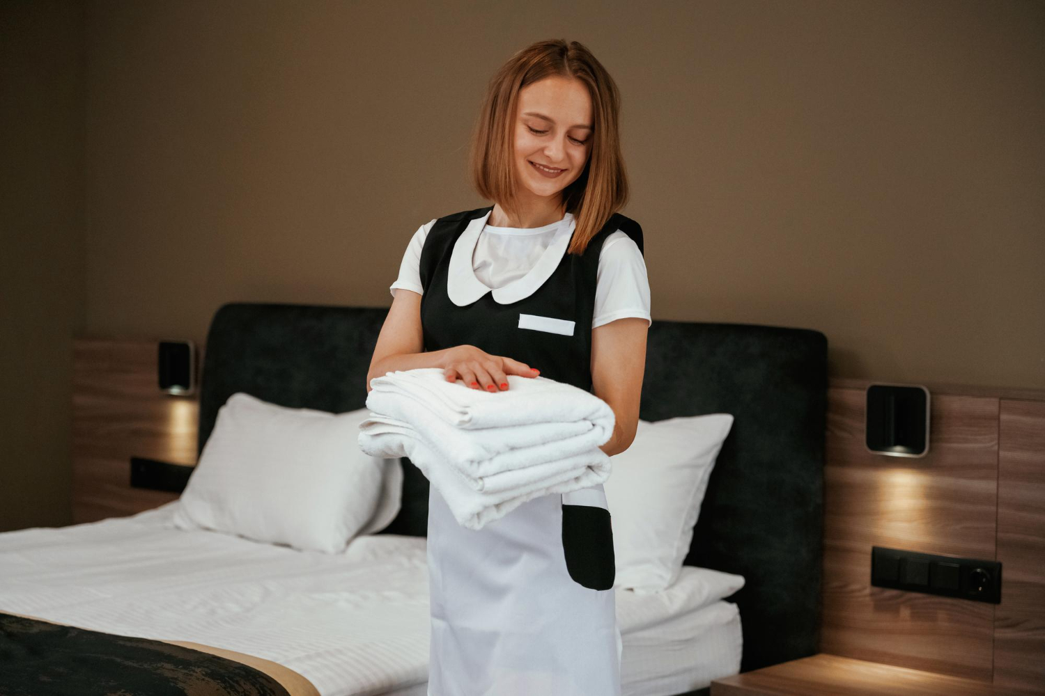 Housekeeper in uniform holding a stack of folded white towels in a hotel room.