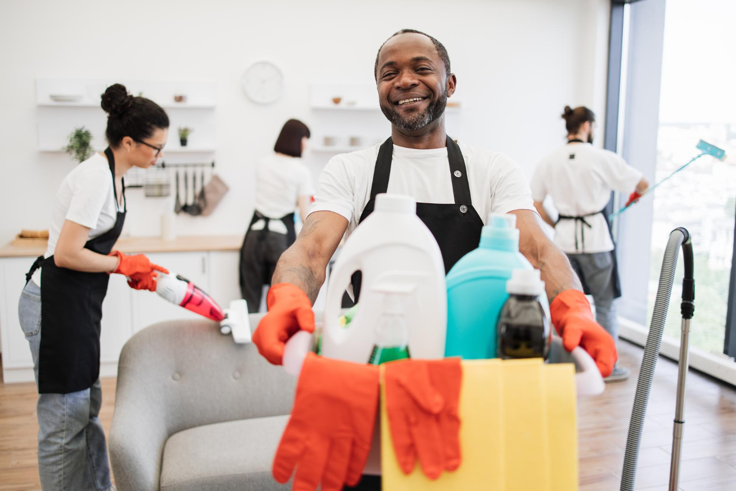Smiling man in gloves holding cleaning supplies with three people cleaning a room in the background.