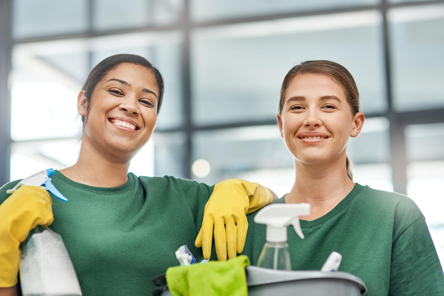 Two smiling women in green shirts and yellow gloves holding cleaning supplies inside a modern building.